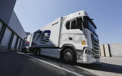 Un camion blanc Scania 770S avec une remorque, garé devant un bâtiment industriel sous un ciel bleu clair.