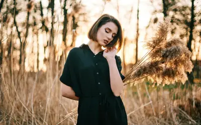 Młoda kobieta w czarnej sukience pozuje na tle zachodzącego słońca i trzcin. Idealna inspiracja na kurs fotografii portretowej.