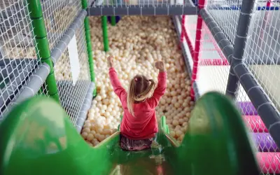 Radosna dziewczynka z uniesionymi rękami zjeżdża po zielonej zjeżdżalni do basenu z piłeczkami w sali zabaw dla niemowląt.