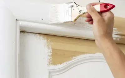 Une main applique de la peinture blanche sur une porte intérieure en bois. Le travail de peinture est en cours, révélant le bois naturel sous la couche blanche.