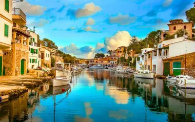 Un pintoresco pueblo pesquero con casas blancas y verdes, barcos amarrados en un canal de aguas cristalinas y un cielo azul con nubes esponjosas.