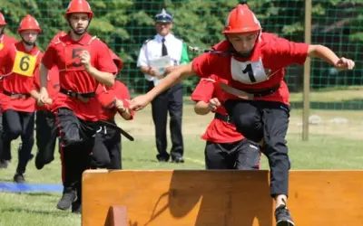 Młodzi strażacy w czerwonych kaskach i strojach biorą udział w zawodach strażackich, pokonując przeszkody.