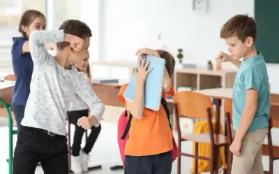 Chłopiec zasłania się zeszytem, podczas gdy inni uczniowie w klasie wydają się być zaangażowani w bullying.