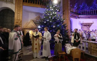 Pasterka w kościele w Zakopanem. Ksiądz czyta z księgi przy choince, obok ministranci i wierni w tradycyjnych strojach.
