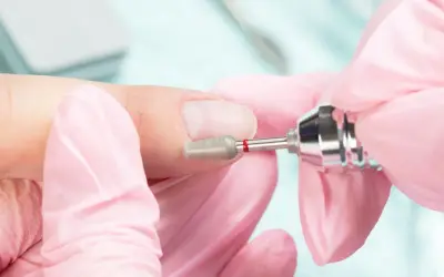 Dłonie w różowych rękawiczkach wykonują manicure frezarką. Przygotowanie paznokci do porodu, by były schludne i bezpieczne.