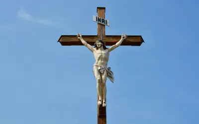Figurka Jezusa na drewnianym krzyżu z tabliczką INRI na tle błękitnego nieba.