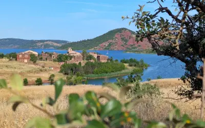 Vue sur le lac du Salagou, avec ses eaux bleues, ses collines rouges et les ruines d'un village. Un voilier navigue paisiblement.