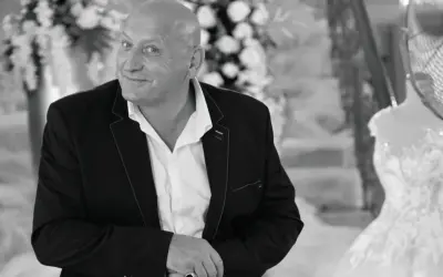 Jacek, znany jako królowe życia, w eleganckim garniturze, z uśmiechem spogląda w obiektyw. W tle widać suknię ślubną.