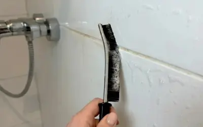 Hand reinigt Duschfugen mit einer Bürste und Schaum.
