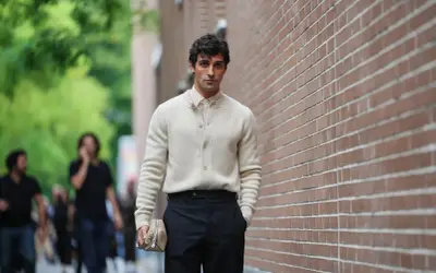 Hombre con suéter crema y pantalón negro, bolso de mano beige. Un ejemplo de las últimas tendencias moda hombre.