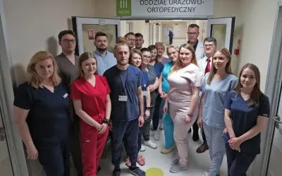 Grupa lekarzy i pielęgniarek w uniformach stoi w korytarzu szpitalnym pod znakiem "Oddział Urazowo-Ortopedyczny".