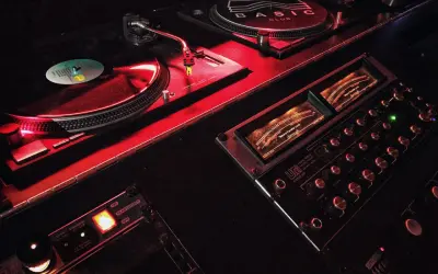 Giradischi e mixer in un locale notturno, illuminati da luci rosse. Il logo "BASIC CLUB" è visibile su uno dei giradischi.