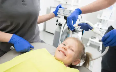 Uśmiechnięta dziewczynka na fotelu dentystycznym, stomatolog przygotowuje sprzęt.