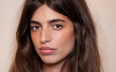 Portrait d'une jeune femme aux longs cheveux bruns. Son maquillage met en valeur ses yeux et ses lèvres, avec un soin particulier apporté à son sourcil poil à poil.