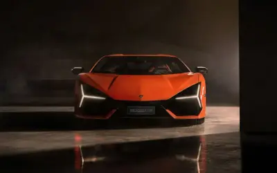 Pomarańczowy Lamborghini Revuelto z ostrymi światłami Y, stojący na błyszczącej podłodze w ciemnym pomieszczeniu.
