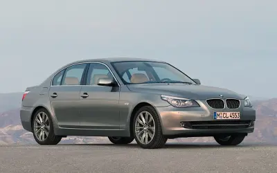 Szary sedan BMW 5 Series (E60) z jasnym wnętrzem stoi na pustej drodze, z górami w tle.