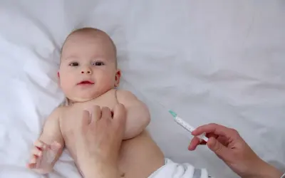 Małe dziecko leży na białym tle, a ręka pielęgniarki przygotowuje szczepienia noworodka w szpitalu.