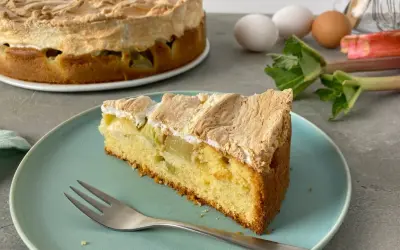 Ein saftiges Stück Rhabarber Baiser Kuchen mit einer luftigen Baiserhaube liegt auf einem blauen Teller. Im Hintergrund ein ganzer Kuchen und frischer Rhabarber.