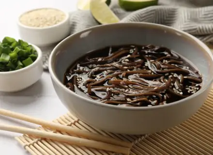 Miseczka z makaronem soba w sosie, posypana sezamem. Obok miseczki z posiekanym szczypiorkiem i sezamem. Zdrowy posiłek, mało makaron soba kcal.