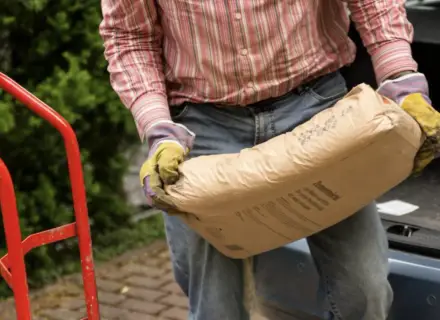 Mężczyzna w rękawicach niesie worek cementu. Zastanawia się, ile piasku na 25 kg cementu będzie potrzebne do budowy.