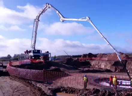 Ogromna pompa do betonu pracuje na budowie, dostarczając materiał do wykopu. Pracownicy w kamizelkach obserwują proces.