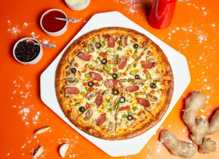 Pizza z kiełbasą, pieczarkami i oliwkami na pomarańczowym tle, obok sosy i imbir.