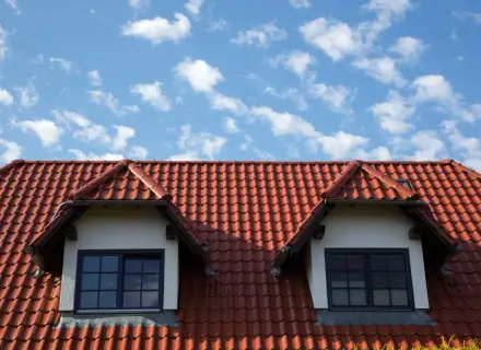 Czerwony dach z dachówki ceramicznej, z dwoma lukarnami i niebieskim niebem w chmurach.