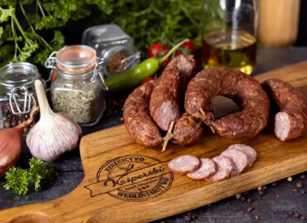 Na drewnianej desce leży pyszna, wędzona kiełbasa jałowcowa, obok czosnek, cebula i przyprawy.