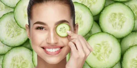 Una mujer sonríe mientras se coloca una rodaja de pepino en el ojo, rodeada de más rodajas de pepino.