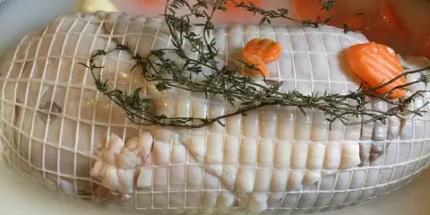 Préparation de la cuisson tête de veau cocotte. Le veau est ficelé, entouré de carottes et de thym dans un bouillon.