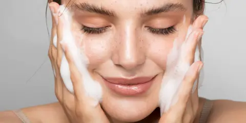 Donna con gli occhi chiusi mentre si lava il viso con un detergente schiumoso. Un gesto delicato per capire come si usa il detergente viso.