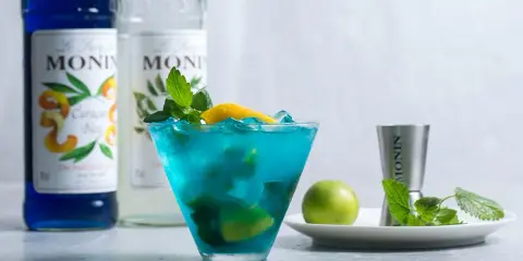 Orzeźwiający drink z syropem blue curacao smak, ozdobiony miętą i cytryną. Obok butelki syropu Monin.
