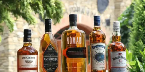 Pięć butelek szkockiej whisky: Edradour, Antiquary, Nomad, Claymore i Whyte & Mackay, stojących na drewnianym stole.