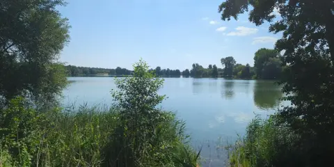 Spokojne jezioro Małe Żnińskie otoczone zielenią, z błękitnym niebem i białymi chmurami odbijającymi się w wodzie.