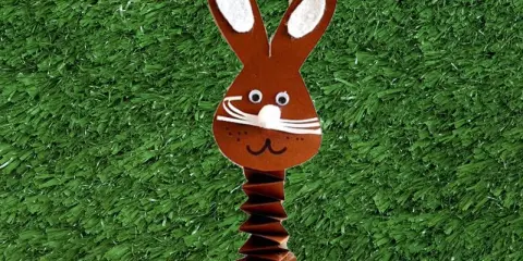 Brauner Papierhase mit Ziehharmonika-Körper, ein süßes Beispiel für Osterhasen basteln, auf grünem Kunstrasen mit bunten Eiern.
