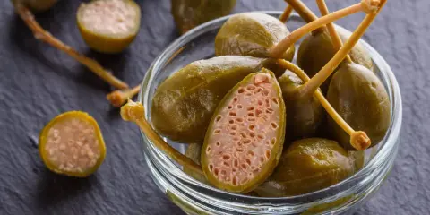 Kapary w szklanej miseczce, jeden przekrojony, na ciemnym tle.