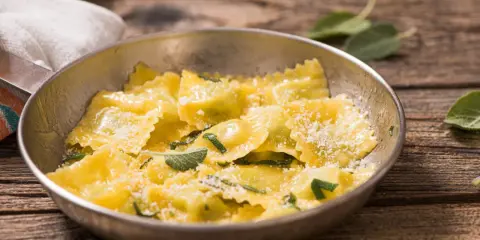 Ravioli ricotta e spinaci dorati, conditi con parmigiano e foglie di salvia fresca, serviti in una ciotola metallica su un tavolo di legno rustico.
