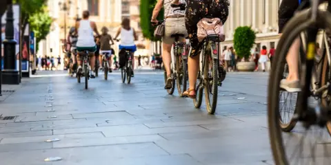 Grupa osób jedzie rowerem po chodniku w słoneczny dzień.