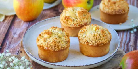 Trois muffins pomme amande dorés avec un crumble croustillant, entourés de pommes fraîches sur une table en bois.