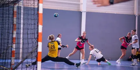 Piłka leci w stronę bramki, gdzie bramkarka w żółtym stroju broni. Zawodniczki w różowych i białych strojach walczą o piłkę na bocznej części boiska.