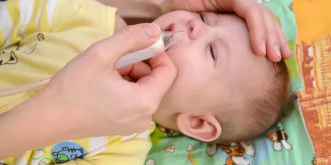 Mani che usano un aspiratore nasale per un neonato con raffreddore. Il piccolo indossa una tutina gialla con disegni di animali.