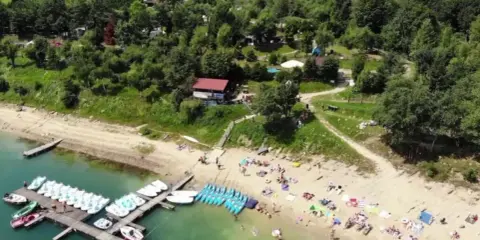 Polańczyk plaża tętni życiem. Ludzie odpoczywają na piasku, a na wodzie czekają rowery wodne i kajaki.