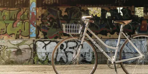 Biały rower z koszykiem, idealny jako rower miejski czy trekkingowy, stoi na tle ściany pokrytej kolorowym graffiti.
