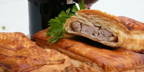 Pâté en croûte, une spécialité lorraine savoureuse, avec une garniture de viande visible. Une bouteille de vin rouge et du persil complètent la scène.
