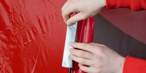 Hände flicken ein rotes Zelt mit silbernem Klebeband.
