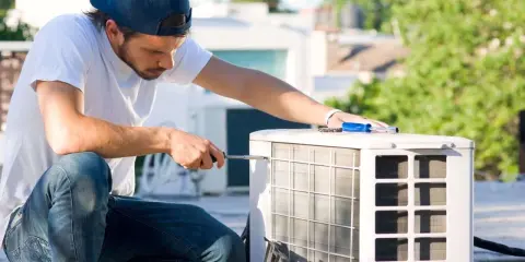 Un technicien effectue l'entretien d'une pompe à chaleur sur un toit, utilisant un tournevis pour ajuster l'unité extérieure.