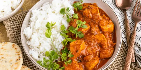 Ein Teller mit dampfendem Reis und cremigem Chicken Tikka Masala, garniert mit frischem Koriander. Dazu Naan-Brot und ein Löffel.