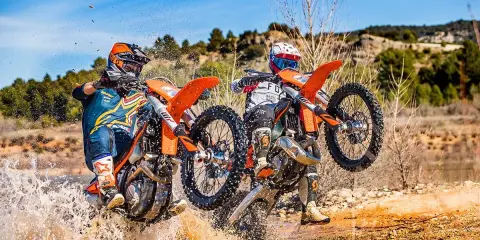 Dos pilotos de enduro compiten en un terreno embarrado, uno con motor 2t y otro 4t, salpicando agua.