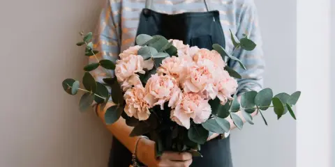 En bukett med ljusrosa nejlikor och eukalyptusblad hålls av en person i förkläde.