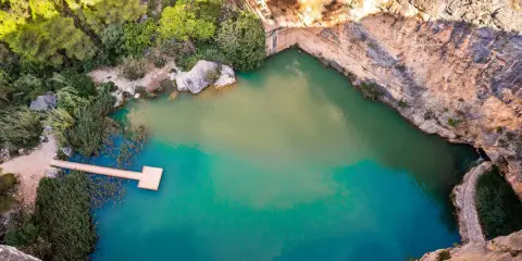 Un **charco azul** en Chiva, rodeado de rocas y vegetación. Un muelle de madera se adentra en sus aguas turquesas.